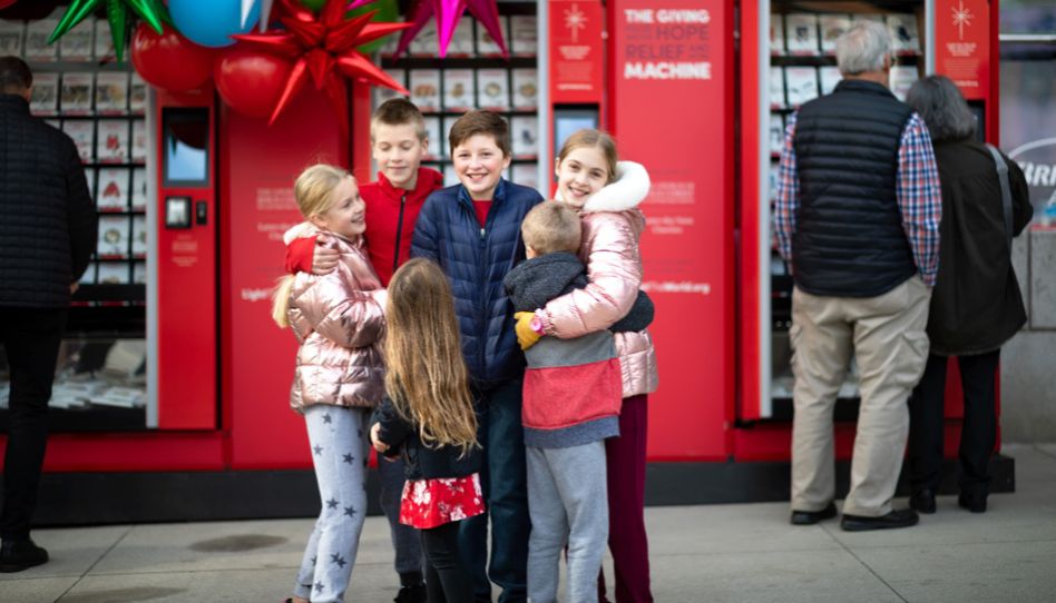 Kinder und Familien stehen fröhlich vor einer Giving Machine der Kirche Jesu Christi und erleben Freude am Geben.