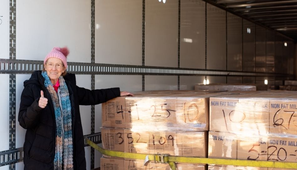 Eine ältere Frau steht lächelnd und mit einem hochgestrecktem Daumen an einer Ladung mit Paketen.