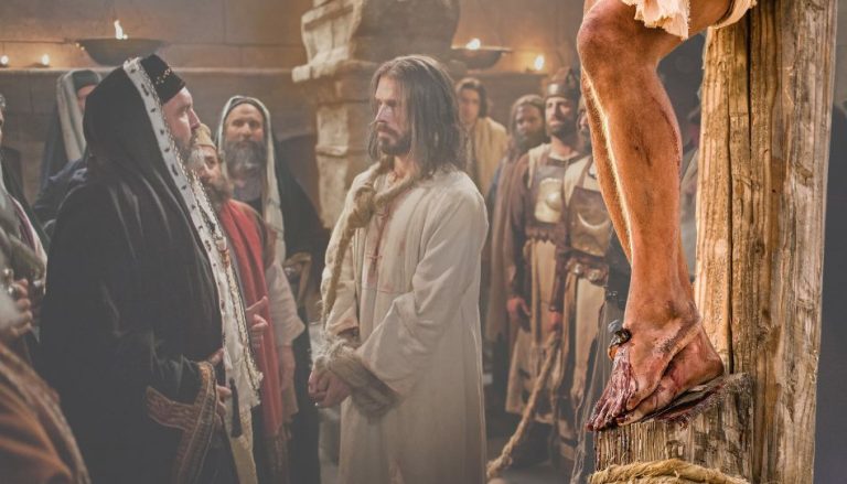 Im Vordergrund sieht man eine Nahaufnahme von Jesus ans Kreuz genagelte Füße. Im Hintergrund wird er (gefesselt) von Kaiphas ausgefragt.