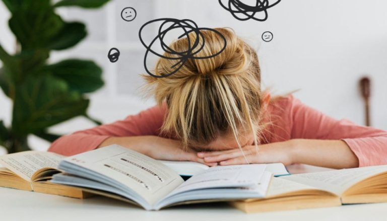 Ein Tisch mit einigen geöffneten Büchern. Ein Mädchen, scheinbar überfordert, legt ihren Kopf auf ihre Hände, die auf den Büchern sind.